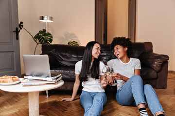 Brunette girls are having fun, clinking glasses of champagne and sitting on floor