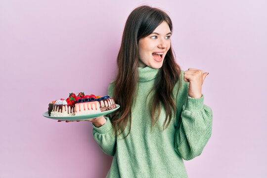 Young beautiful caucasian girl holding cake slices pointing thumb up to the side smiling happy with open mouth