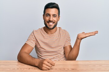 Handsome hispanic man wearing casual clothes sitting on the table smiling cheerful presenting and...