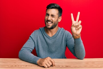 Handsome hispanic man wearing casual style sitting on the table smiling with happy face winking at...