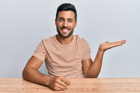 Handsome Hispanic Man Wearing Casual Clothes Sitting On The Table Smiling Cheerful Presenting And Pointing With Palm Of Hand Looking At The Camera.