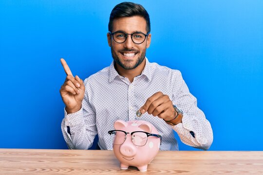 Handsome hispanic man holding piggy bank with glasses smiling happy pointing with hand and finger to the side