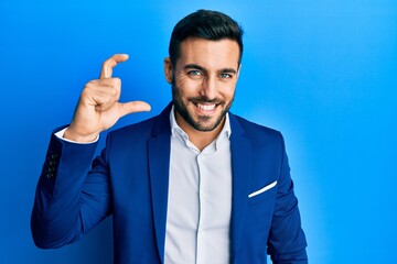 Young hispanic businessman wearing business jacket smiling and confident gesturing with hand doing small size sign with fingers looking and the camera. measure concept.