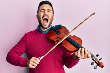 Young hispanic musician man playing violin angry and mad screaming frustrated and furious, shouting with anger looking up. © Krakenimages.com