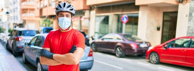 Fototapeta na wymiar Young delivery man wearing bike helmet and coronavirus protection medical mask standing at town street.