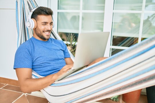 Young hispanic man relaxed working using laptop and headphones lying on the hammock at terrace.