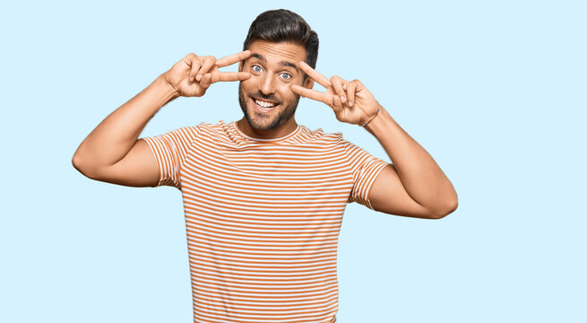 Handsome hispanic man wearing casual clothes doing peace symbol with fingers over face, smiling cheerful showing victory