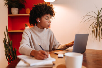 Fototapeta na wymiar Dark-skinned girl in glasses and headphones looks at laptop screen and writes notes in notebook