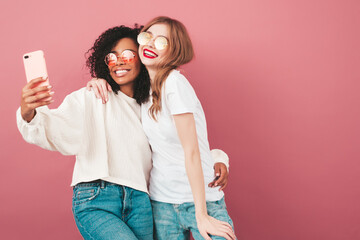 Two young beautiful smiling international hipster female in trendy summer jeans clothes. Sexy carefree women posing near pink wall in studio. Positive models having fun. They taking selfie photos