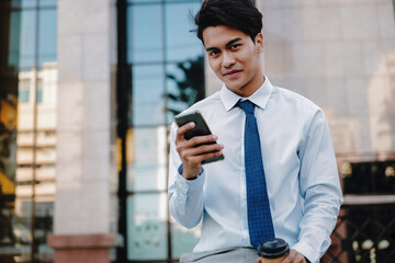 Smiling Young Asian Businessman Using Mobile Phone in the City. Looking at the Camera