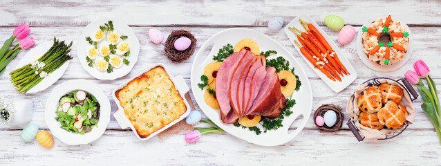Classic Easter ham dinner. Top down view table scene on a white wood banner background. Ham, scalloped potatoes, eggs, hot cross buns, carrot cake and vegetables.