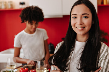 Asian brunette woman in striped top is smiling, looking into camera. Curly dark-skinned lady cooks salad