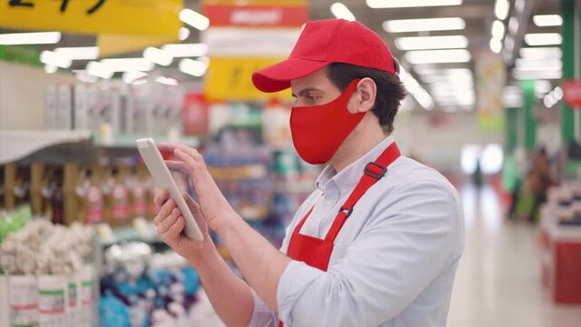 Caucasian male store employee in red face mask and uniform standing in supermarket and typing on tablet computer. Seller working in delivery service during epidemic covid-19, coronavirus pandemic