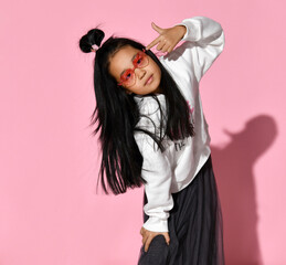 Fashionable portrait of a little Korean model girl who is professionally posing on a pink background. Girl is wearing a black skirt, white sweater, sneakers and red glasses. Kids fashion concept.