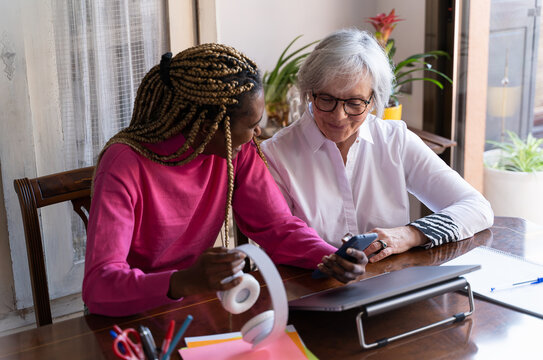 Young African Woman Smiling As She Teaches An Older Woman How To Use A Mobile Phone Indoors