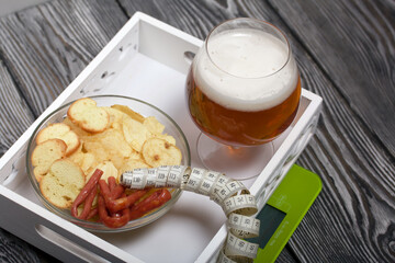 A glass of beer, sausages, chips and croutons. Scales and measuring tape are nearby. On pine boards. A day without diets.