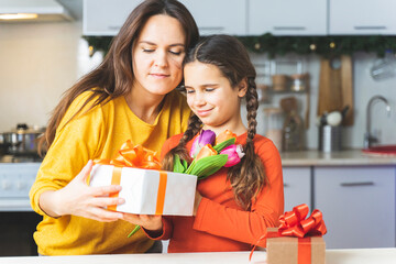 A cute girl congratulates her mother in the kitchen of the house on her birthday. Happy family: kid gives his mom a gift and kisses