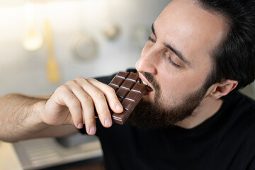 A man emotionally eats chocolate bars at home in the kitchen. 