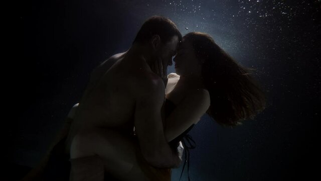 brunette lady and brawny man are hugging inside water of pool, underwater view