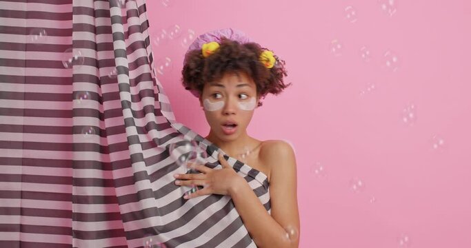 Embarrassed shocked Afro American woman reacts puzzled as someone came in into bathroom hides naked behinds shower curtain undergoes daily hygiene procedures poses around foam bubbles around