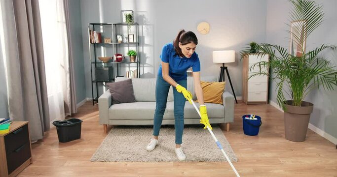 Beautiful happy young Caucasian woman in yellow gloves washing floor at home in living room. Pretty female working mopping floor in apartment doing domestic work, housekeeper concept