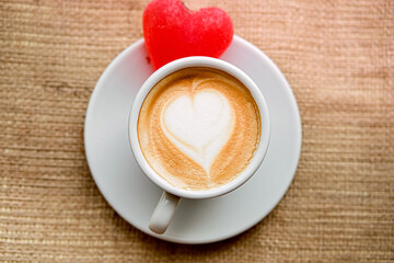 one white cup on a saucer with  coffee and marmalade heart stands on a windowsill on burlap fabric, loneliness.  drink with milk, with foam heart, top view.