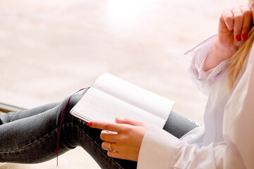 Beautiful sexy girl sits on a windowsill with a notebook and thinks. Details close up. A lonely woman in a man's shirt reads a book, keeps notes in a notebook.