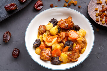 A dish of Jewish cuisine sweet tsimes with dates, carrots and turkey meat in a white plate on a concrete background near raisins and dates.