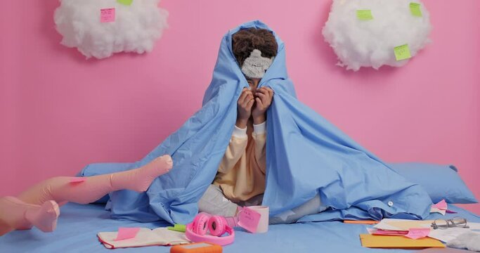 Scared Afro American woman hides behind blue blanket poses on bed studies from home has inflated doll for love stimulation feels lonely and insecure dressed in nightwear. Distance education.