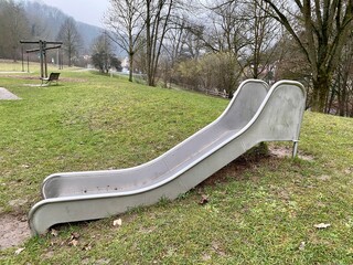Stahl - Rutsche steht auf abgelegenen Kinderspielplatz, hinten Holzkarussell kahle Bäume und Wald...