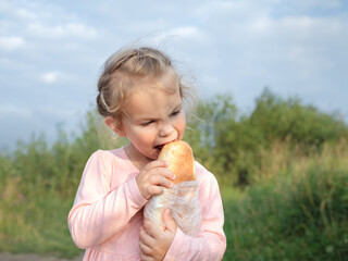 Little toddler girl holding big loaf of bread. Kid biting and eating healthy bread, outdoors. Hungry kid.