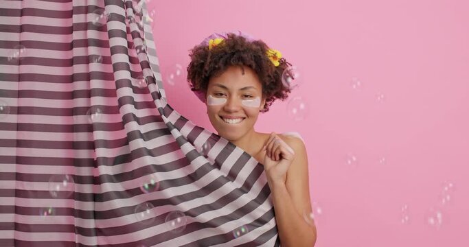 Overjoyed upbeat Afro American woman has fun while taking shower moves energetically hides naked body with curtain relaxes in bathroom cares about skin and body isolated over pink background