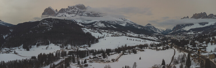 Winter aerial drone panorama of Cortina d'Ampezzo in cold and cloudy day with visible city in the background.