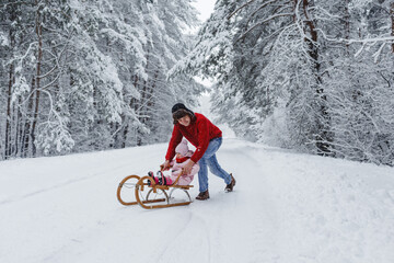 A young father rides a wooden sled with his adorable daughter in a snow-covered forest.