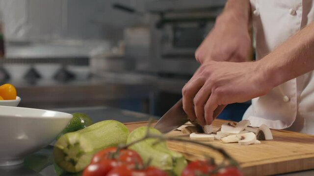 Midsection close-up of unrecognizable male chef in white uniform dicing raw mushrooms on wooden cutting board at restaurant kitchen