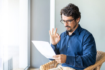 Man wearing glasses on a chair holding paper document. Male portrait reading contract files and...