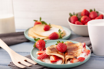Imagen de un plato de crepes con trozos de fresas y nata y otro detras con azucar glass sobre un fondo claro y un cuenco de fresas con una taza y una botella de leche.