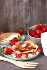 Vista en vertical de un plato de crepes con trozos de fresa y nata, otro plato de crepes con azúcar glass en el fondo y un cuenco de fresas