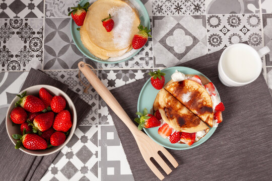 Vista cenital de dos platos de crepes, uno de ellos con nata y trozos de fresa y otro con az&uacute;car glass. Un cuenco de fresas a la izquierda y una taza de leche a la derecha.