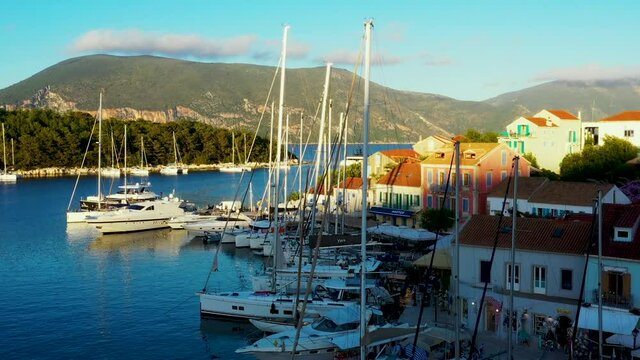 Drone taking off over the beautiful marina and town of Fiskardo in Greece. There are a lot of boats in the marina. It is a beautiful evening around sunset. There are mountains in the background. 1080p