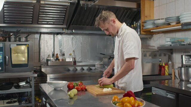Slow-motion medium side-view shot of professional caucasian chef cooking healthy dish, slicing some fresh organic zucchini on wooden cutting board in restaurant kitchen