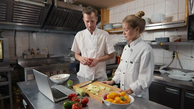 Medium shot of skilled chef teaching young female cook basics of cutting with chef knife, showing video tutorial on laptop during practice at restaurant kitchen