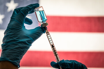 Front Line Worker Holding Syringe and Vial Filled with Coronavirus Vaccine or Medicine Silhouetted Against American Flag