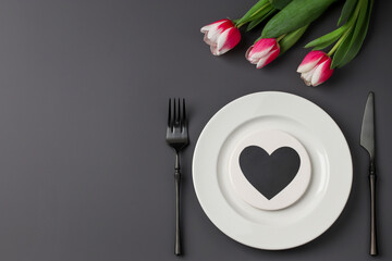 Creative table setting with hearts on a white plate, black fork and knife, tulips on a dark background. Valentine's Day, Wedding Day, Birthday, Women's Day and Mother's Day. Flat lay. Copy space