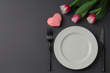 Creative table setting with hearts, white plate, black fork and knife, tulips on a dark background. Valentine's Day, Wedding Day, Birthday, Women's Day and Mother's Day. Flat lay. Copy space