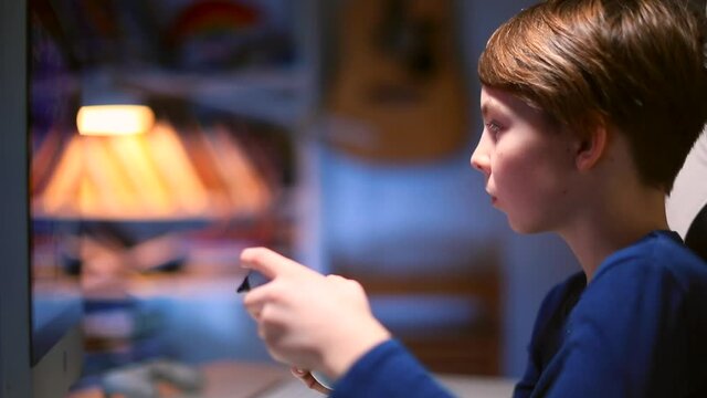 Focus of attention of a teenager boy on a monitor in a video game ps4. In his hands, the joystick controls the application without a wire. Evening room.