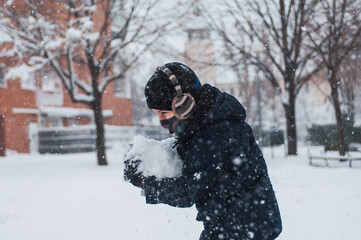 Fototapeta na wymiar boy carrying a large snowball during a snowfall wearing a mask against covid winter hat, gloves and earmuffs. he is wearing a blue coat. Lifestyle