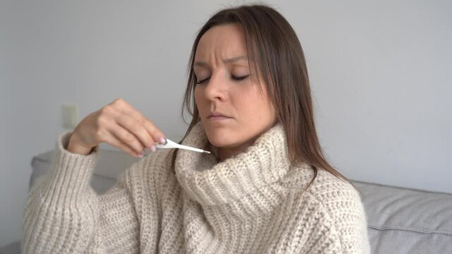 Sad sick young woman with fever measuring temperature with medical thermometer at home