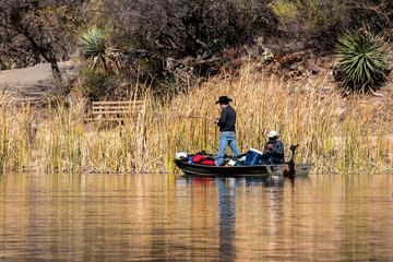 Fototapeta na wymiar Two people fish from a small boat at Patagonia Lake State Park, Arizona. Both men are wearing face coverings. Their boat is overfilled with gear. Concepts of outdoor recreation, fishing, state parks