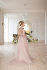 Fototapeta premium A beautiful young bride in a beige vintage dress with a bouquet of yellow and white flowers on the background of a classic light interior with spring floral decor. Elegant indoor wedding ceremony.